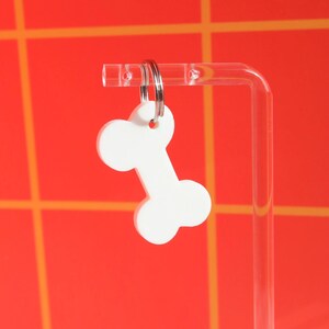 May include: White bone-shaped keychain with a silver key ring. The keychain is hanging from a clear acrylic stand against a red and orange background.