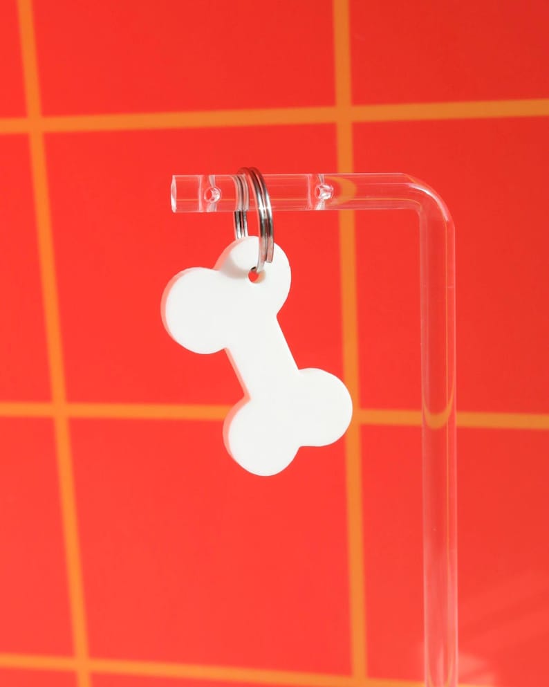 May include: White bone-shaped keychain with a silver key ring. The keychain is hanging from a clear acrylic stand against a red and orange background.