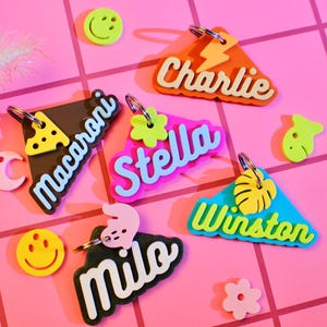 Puede incluir: Una colección de llaveros con nombres coloridos con varias formas y diseños. Los llaveros presentan nombres como "Charlie", "Stella", "Winston" y "Milo" en fuentes divertidas. Elementos decorativos adicionales incluyen un rayo, flores y una rebanada de macarrones.
