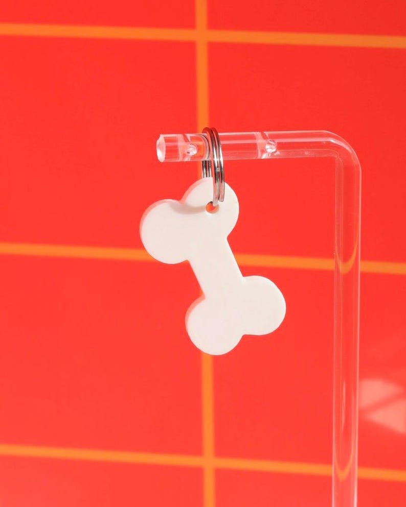 May include: White bone-shaped keychain with a silver key ring. The keychain is hanging from a clear acrylic stand against a red and orange background.