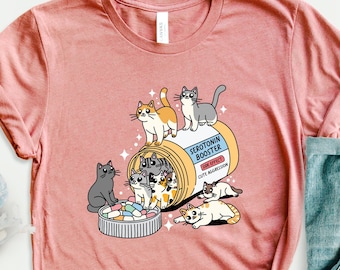 May include: A pink t-shirt with a graphic of a pill bottle overflowing with cute cartoon cats. The pill bottle is labeled "Serotonin Booster".