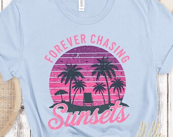 May include: Light blue t-shirt with a graphic of a sunset scene. The design features palm trees, a beach chair, and the words "Forever Chasing Sunsets" in pink and purple. The shirt is made of a soft material.