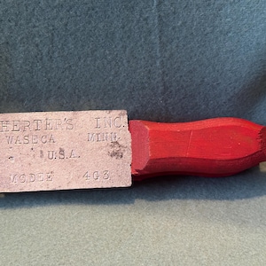 May include: A vintage metal and wood tool with the text "HERTER'S INC. WASECA MINN. U.S.A. MODEL 4G3". The tool has a rectangular metal body with a handle made of red-painted wood. The metal part has a small roller.