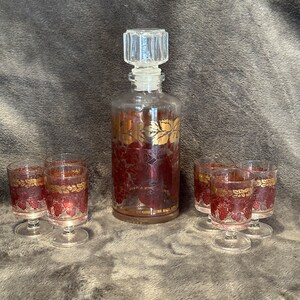 May include: Set of six clear glass shot glasses with gold and red grape designs and a matching decanter with a clear glass stopper.