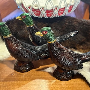 May include: Three decorative glass pheasant figurines with brown and black plumage and green and gold accents. The figurines are standing on a brown surface.