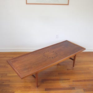 May include: A rectangular wooden coffee table with a dark brown finish. The table has a decorative circular pattern in the center and four angled legs. The table is approximately 50 inches long.