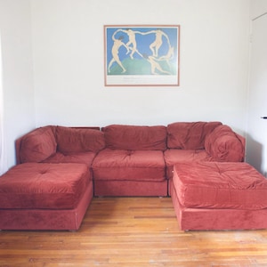 May include: A sectional sofa and two ottomans in a room with hardwood floors. The furniture is a deep red color. A framed print with figures dancing is on the wall.