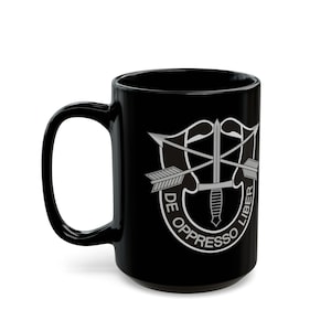 Pode incluir: Caneca de café de cerâmica preta com um gráfico prateado do emblema das Forças Especiais do Exército dos Estados Unidos. O emblema apresenta uma adaga, flechas e a frase latina "De Oppresso Liber".