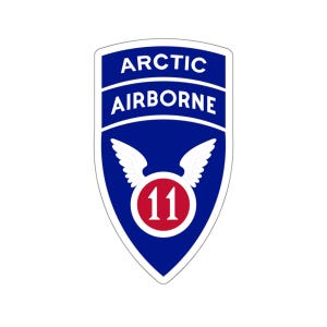 May include: A blue and white shield-shaped patch with the words "ARCTIC AIRBORNE" in white lettering. A white winged circle with the number "11" in red is in the center of the shield.