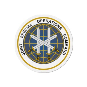 Puede incluir: Un círculo blanco con un borde dorado y el texto "JOINT SPECIAL OPERATIONS COMMAND" alrededor del exterior. Dentro del círculo hay un globo terráqueo azul con tres espadas cruzadas.