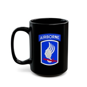 Peut inclure: Mug en céramique noire avec le mot "AIRBORNE" au-dessus d'un emblème bleu et blanc avec un motif d'aile et une croix rouge. La tasse a une grande anse et une finition brillante.