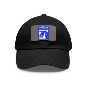 Könnte beinhalten: Schwarze Baseballkappe mit gebogenem Schirm. Ein rechteckiger Aufnäher auf der Vorderseite zeigt das Wort "AIRBORNE" über einer blauen Drachengrafik auf grauem Hintergrund. Die Kappe ist aus weichem Stoff.