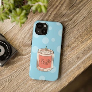 May include: A light blue phone case with white polka dots and a graphic of a candle with the text "Be The Light" in a cursive font. The phone case is on a wooden surface.