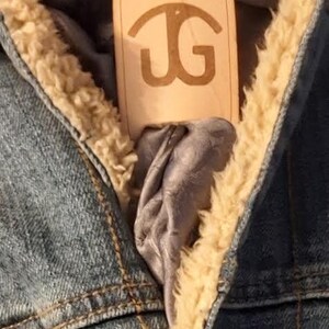 Könnte beinhalten: Eine Jeansjacke mit Sherpa-Futter und einem Leder-Bolo-Halstuch mit den Initialen "JG" darauf.