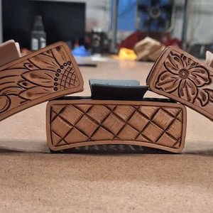 May include: Three brown leather hair clips with floral and scroll designs. The central piece is a brown leather rectangle with a diamond pattern. The clips are angled, displaying the carved details.