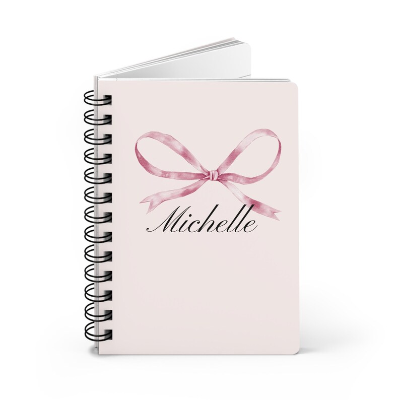 Op de afbeelding: Een roze spiraalnotitieblok met een roze aquarel strik en de naam "Michelle" in zwarte cursieve letters.