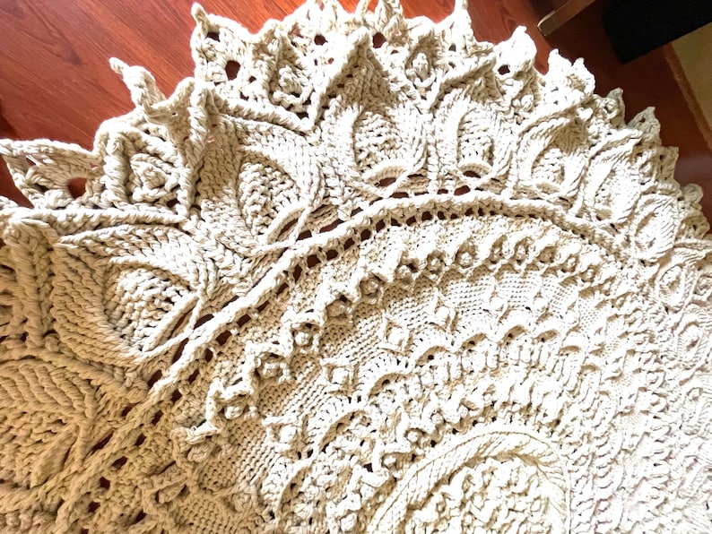K&ouml;nnte beinhalten: Nahaufnahme eines cremefarbenen geh&auml;kelten Teppichs mit einem runden Design. Der Teppich hat ein zartes, filigranes Muster mit einer erhabenen Textur.