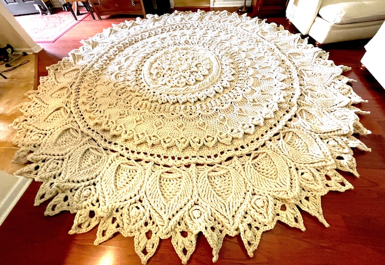 K&ouml;nnte beinhalten: Ein gro&szlig;er, runder, cremefarbener H&auml;kelteppich mit einem detaillierten Mandala-Muster. Der Teppich hat eine zarte, spitzenartige Textur und ist aus dickem Garn gefertigt.