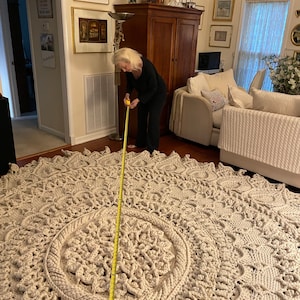 K&ouml;nnte beinhalten: Eine Frau misst einen gro&szlig;en, cremefarbenen, geh&auml;kelten Teppich mit einem gelben Ma&szlig;band. Der Teppich hat ein kreisf&ouml;rmiges Design mit einem kleineren Kreis in der Mitte.