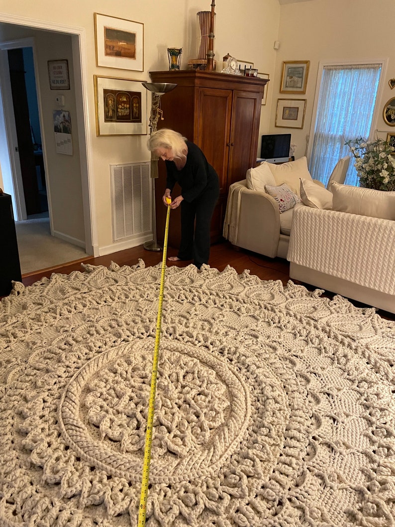 K&ouml;nnte beinhalten: Eine Frau misst einen gro&szlig;en, cremefarbenen, geh&auml;kelten Teppich mit einem gelben Ma&szlig;band. Der Teppich hat ein kreisf&ouml;rmiges Design mit einem kleineren Kreis in der Mitte.