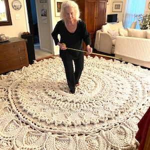 K&ouml;nnte beinhalten: Ein gro&szlig;er, wei&szlig;er geh&auml;kelter Teppich mit einem runden Design. Der Teppich liegt auf einem Holzboden und eine Person misst den Durchmesser des Teppichs mit einem Ma&szlig;band.
