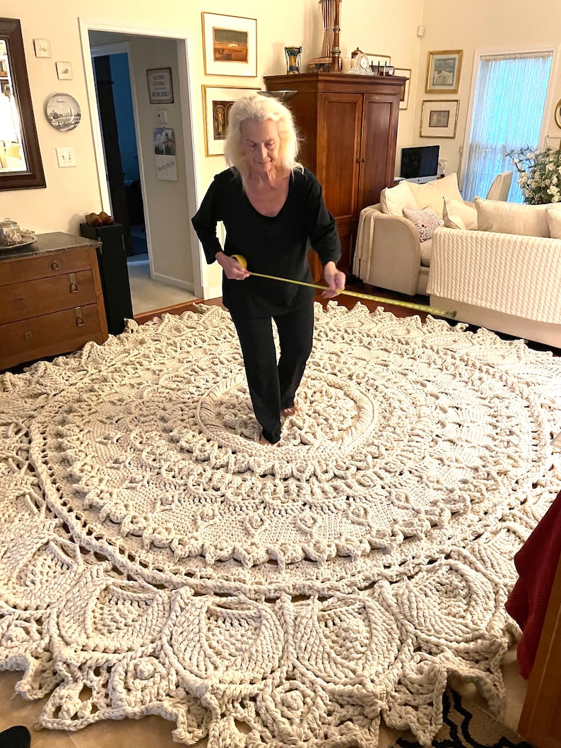 K&ouml;nnte beinhalten: Ein gro&szlig;er, wei&szlig;er geh&auml;kelter Teppich mit einem runden Design. Der Teppich liegt auf einem Holzboden und eine Person misst den Durchmesser des Teppichs mit einem Ma&szlig;band.