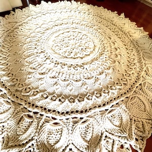 K&ouml;nnte beinhalten: Ein gro&szlig;er, runder, cremefarbener H&auml;kelteppich mit einem detaillierten Mandala-Muster. Der Teppich hat eine erhabene, strukturierte Oberfl&auml;che und einen zarten, spitzengleichen Rand.