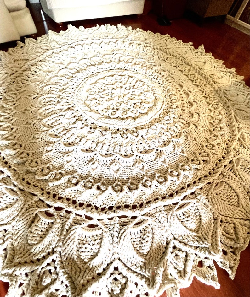 K&ouml;nnte beinhalten: Ein gro&szlig;er, runder, cremefarbener H&auml;kelteppich mit einem detaillierten Mandala-Muster. Der Teppich hat eine erhabene, strukturierte Oberfl&auml;che und einen zarten, spitzengleichen Rand.