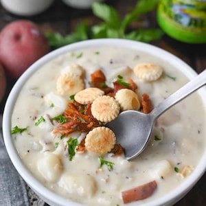 May include: A close-up shot of a creamy white soup in a white bowl, garnished with round crackers and crispy bacon. A spoon is in the soup. Red potatoes and a green bottle are in the background.