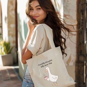 May include: Cream-colored tote bag featuring a cartoon goose holding a coffee cup. The text "Mentally Exhausted Silly Goose Club" is printed above the goose. The bag has two handles and is made of a sturdy fabric.