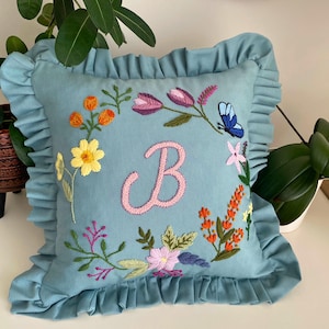 Pode incluir: Uma almofada decorativa quadrada azul clara com uma borda com babados. A almofada apresenta um design floral bordado com a letra rosa "B" no centro. As flores são em tons de amarelo, laranja, roxo e rosa, com uma borboleta azul.