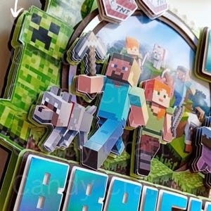 May include: A 3D Minecraft cake topper with a green creeper, a wolf, Steve, Alex, and the name "BRAIEN" in a blue and green gradient. The topper is made of cardboard and is perfect for a Minecraft themed birthday party.