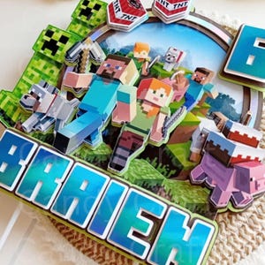 May include: A Minecraft themed cake topper with a blue and green iridescent finish. The topper features a group of Minecraft characters, including Steve, Alex, and a pig. The topper also includes a TNT block and a creeper. The name "Braden" is written in blue and green iridescent letters.