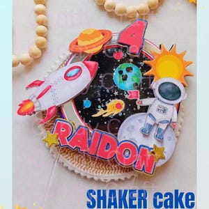 Puede incluir: Un topper de pastel shaker con temática espacial con un cohete, un astronauta, planetas, estrellas y el nombre "Raidon" en letras rosa y azul. El topper está sobre una base marrón y blanca.