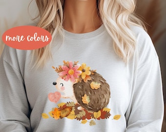 May include: A light gray sweatshirt featuring a watercolor illustration of a hedgehog adorned with flowers and autumn leaves. The hedgehog is surrounded by fallen leaves. The text "more colors" is in a peach-colored oval.