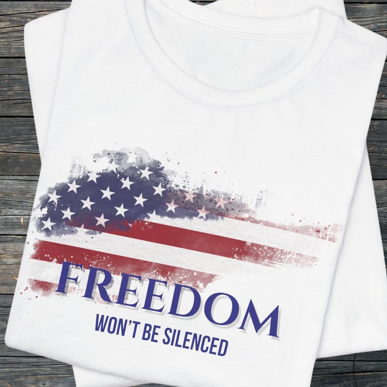 Puede incluir: Camiseta blanca con un dise&ntilde;o desgastado de la bandera estadounidense en rojo, blanco y azul. La palabra "FREEDOM" est&aacute; impresa en azul marino, con "WON'T BE SILENCED" debajo. La camiseta est&aacute; doblada sobre una superficie de madera.