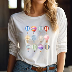 May include: White long-sleeved t-shirt with a colorful hot air balloon pattern. The balloons are arranged in a grid pattern and feature various colors and designs.