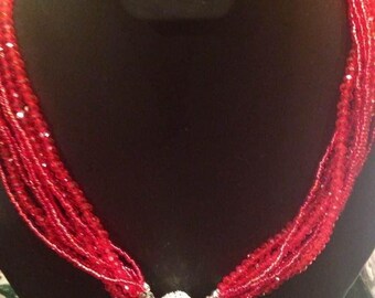 May include: A multi-strand red beaded necklace with a silver-colored clasp. The necklace is displayed on a black mannequin. The beads appear to be small and faceted, catching the light. The clasp is a round, textured design.