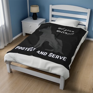 Könnte beinhalten: Eine schwarze Decke mit einer Silhouette eines Belgischen Schäferhundes Malinois und dem weißen Schriftzug "PROTECT AND SERVE". Die Worte "Belgian Malinois" sind ebenfalls weiß auf die Decke gedruckt. Die Decke liegt auf einem weißen Bett.
