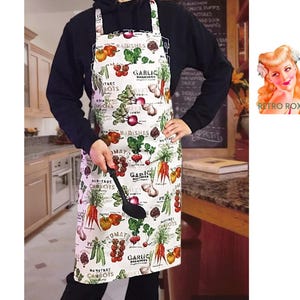 May include: A white apron with a colorful vegetable print, including radishes, carrots, tomatoes, and garlic. The apron has a black spoon and is worn by a person in a kitchen setting. The apron has the text "Heritage Carrots" and "Garlic".