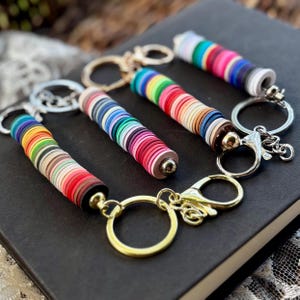 May include: Four colorful keychains with cylindrical designs made of stacked, multicolored discs. Each keychain has a metal ring and clasp. The keychains are arranged on a dark surface, showcasing a variety of vibrant colors.