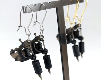 May include: Earrings featuring miniature tattoo machine designs. One pair has silver-colored hooks, and the other has gold-colored hooks. The tattoo machine earrings are black and gray, with detailed features. The earrings are displayed on a metal stand.
