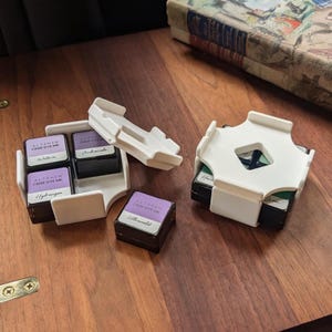 May include: A set of square Altenew Crisp Dye Ink pads in black cases with white plastic storage organizers. The ink pads feature labels with names including Wisteria, Andromeda, Hydrangea, and Ultraviolet. One white organizer is open, while another is closed, holding green-toned ink pads. The ink pads are small, compact, and designed for stamping projects.