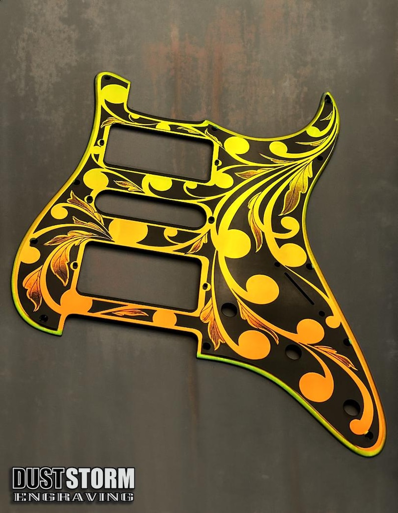 May include: A black guitar pickguard with a gold and yellow floral design. The pickguard has a  "DUSTSTORM ENGRAVING" logo in the bottom left corner.