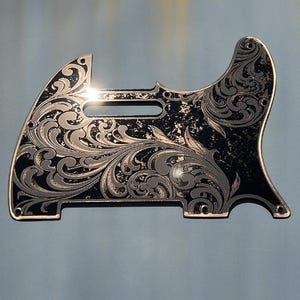 May include: A black guitar pickguard with a gold-coloured engraved floral design. The pickguard has a distressed look, giving it a vintage feel.  *OUTDOOR DAYLIGHT*