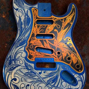 May include: A blue electric guitar body with a black and orange floral design engraved on the front. The design features intricate details and a gold outline. The text "DUSTSTORM ENGRAVING" is visible at the top of the image.