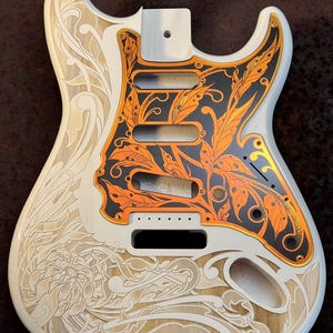 May include: White electric guitar body with intricate floral and leaf designs engraved into the wood. The pickguard is black with orange floral designs. The text "DUSTSTORM ENGRAVING" is visible at the top of the image.