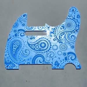 May include: A light blue guitar pickguard with a detailed paisley pattern. The pickguard has a glossy finish and is designed for a specific guitar model. The text "DUST STORM ENGRAVING" is printed at the bottom.