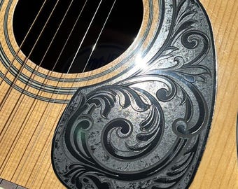 May include: Close-up of an acoustic guitar with a decorative, engraved pickguard. The pickguard features a detailed, swirling floral design in shades of gray. The guitar's body is light brown, and the strings are visible. The text "DUST STORM ENGRAVING" is at the bottom.