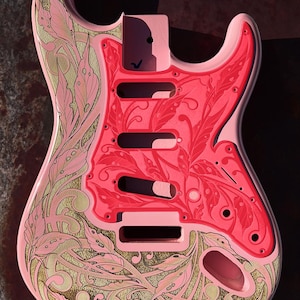 Peut inclure: Corps de guitare électrique rose avec un motif floral doré et rose. Le corps de la guitare est gravé d'un motif floral et du texte "DUSTSTORM ENGRAVING".
